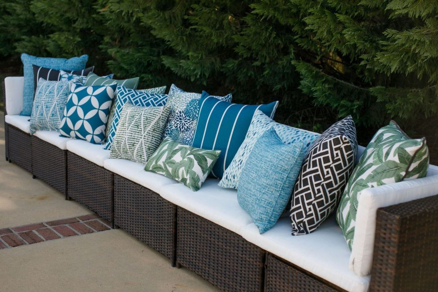 Outdoor pillow covers from various color collections from turquoise & teal to black in patterns from striped, geometric to tropical.  All pillows are stuffed and sitting on a white sofa situated on a patio. 